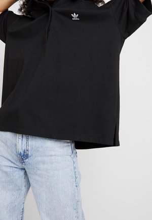 Osoba v černé polokošili Adidas s logem uprostřed na hrudi, v kombinaci s světle modrými džínami, s rukama zdviženýma mimo rám.