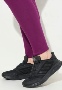 Person i svarta Adidas löparskor och lila leggings, stående mot en vit bakgrund.