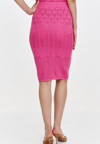 Szydełkowana spódnica ołówkowa w jasnoróżowym kolorze z teksturowanymi wzorami i dopasowanym krojem. Dołek spódnicy znajduje się tuż poniżej kolan. Noszona z beżowymi klapkami.