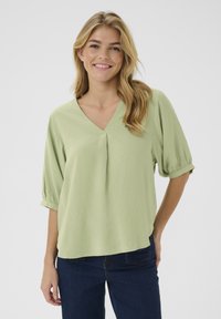 Top verde chiaro a maniche corte con scollatura a V e dettagli plissettati. Realizzato in tessuto morbido con una vestibilità rilassata, abbinato a jeans blu scuro.