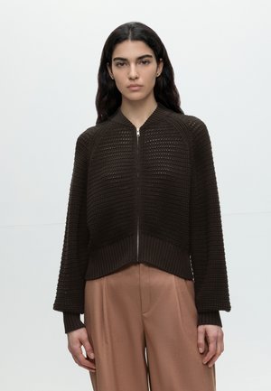 Jonge vrouw met lang donker haar, die een donkerbruine gestructureerde trui met rits en lichtbruine hoge taille broek draagt, staand tegen een effen achtergrond.