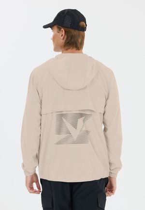 Mann trägt beigen Kapuzenjacke mit abstraktem Logo auf dem Rücken und schwarze Mütze, blickt mit dem Rücken zum einfarbigen Hintergrund.