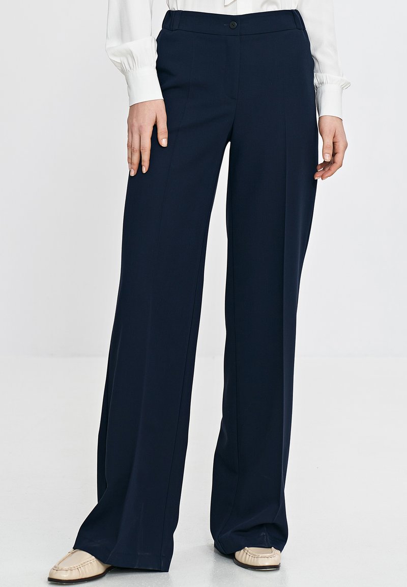 Granatowe spodnie o szerokich nogawkach wykonane z gładkiego materiału, z fałdami z przodu. Posiadają zapięcie na guzik i kieszenie, zestawione z jasno kolorowymi butami.