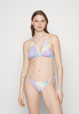 Νεαρή γυναίκα που φοράει ένα παστέλ μπικίνι με σχέδιο tie-dye σε τριγωνικό σχήμα και μπικίνι με κορδόνια στο πλάι, στέκεται μπροστά από ένα απλό ανοιχτό φόντο.