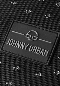 Fekete textúrájú szövet, amelyen szórványosan fényes részletek találhatók, valamint egy téglalap alakú gumicímke, amelyen a "JOHNNY URBAN" felirat nagybetűkkel van gravírozva.