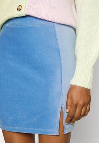 Saia mini em veludo cotelê em gradiente de azul, com fenda lateral. O tecido apresenta uma textura ribeteada. Combinada com um suéter de malha em tom pastel.
