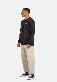 Sudadera negra con cuello redondo y pequeño logo, combinada con pantalones holgados de un tono beige claro y zapatillas de alto corte en azul marino.