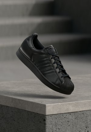 Sort lædersneaker med struktureret gummitåkappe, flade snørebånd og perforerede sidestreger, placeret over en betonoverflade.