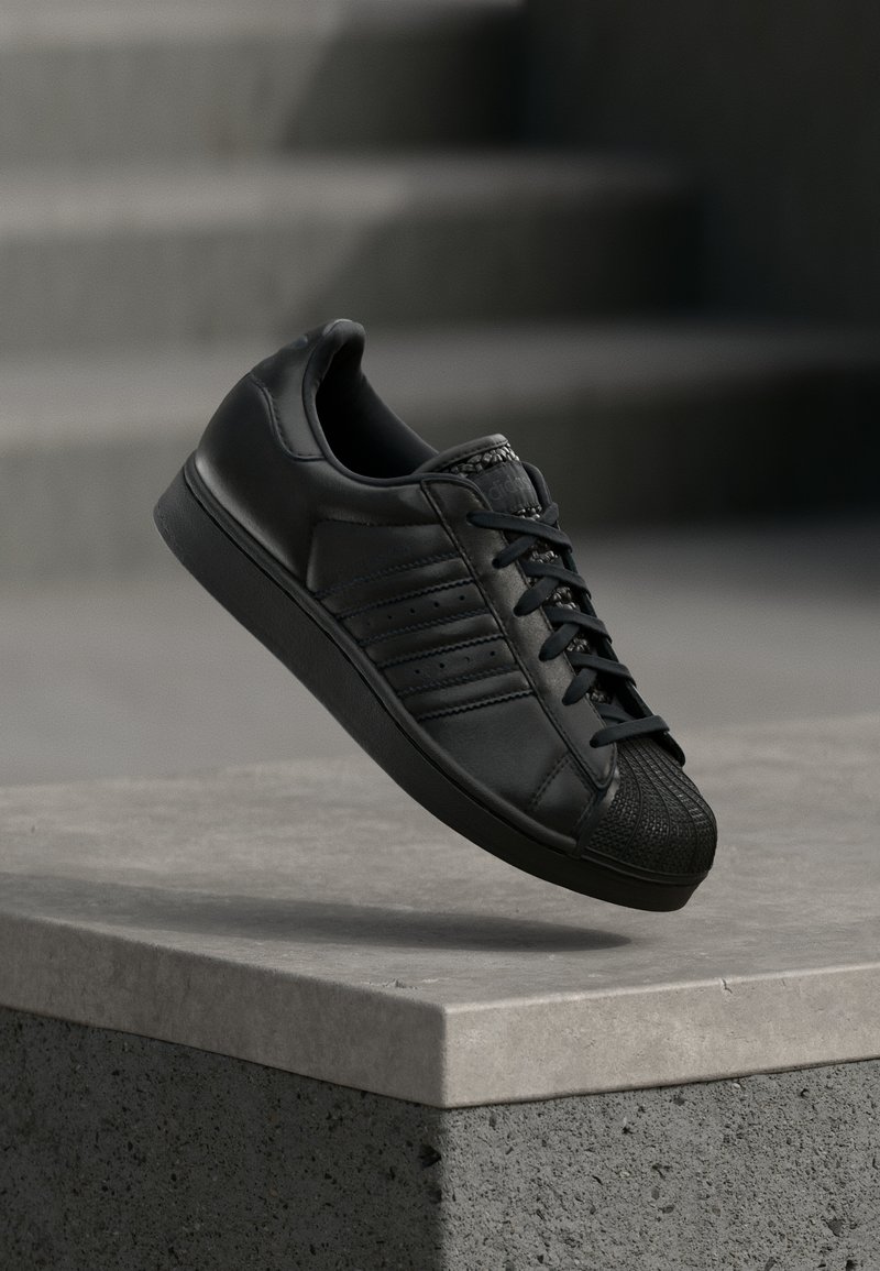 Baskets en cuir noir avec bout en caoutchouc texturé, lacets plats, et bandes latérales perforées, posées sur une surface en béton.