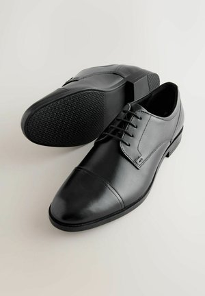 Ein Paar schwarze Leder-Hausschuhe mit Schnürsenkeln, einer zeigt eine glatte Sohle mit strukturiertem Griff, auf hellem Hintergrund.