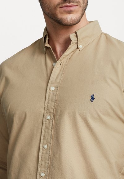 Beige skjorta med knappar i texturerad bomull och klassisk krage, med vita knappar och en liten blå broderad logotyp på bröstet.