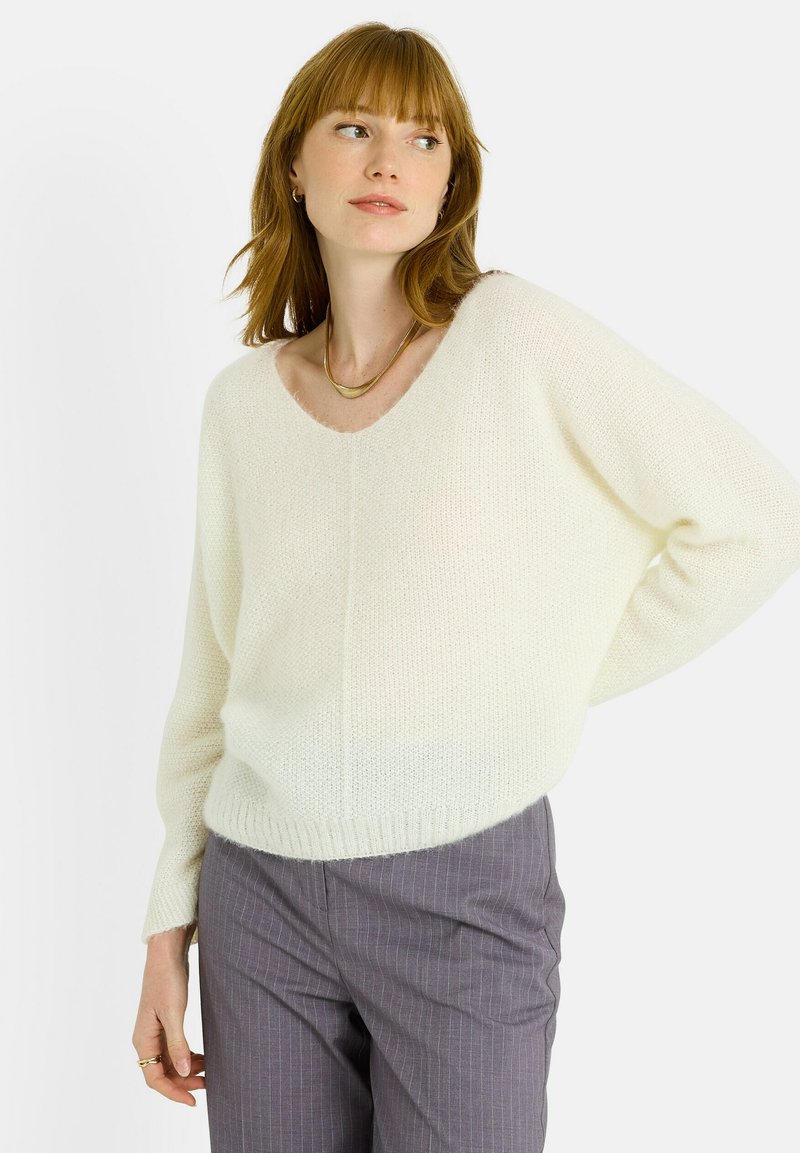 Suéter de punto color crema claro con escote en V, ajuste holgado y dobladillo acanalado. Textura suave y hombros caídos, llevado sobre pantalones con estampado.