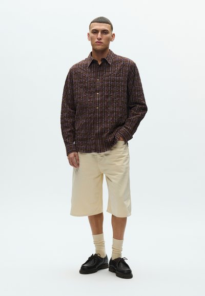 Καφέ πουκάμισο με καρό κουμπιά και υφή ύφασμα, σορτς εκρού μέχρι το γόνατο, ανοιχτό μπεζ κάλτσες και μαύρα δερμάτινα παπούτσια με κορδόνια.