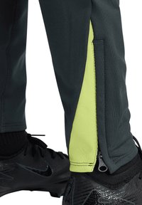 Sporthose aus dunkelgrünem Stoff mit einer leuchtend gelben Seitenblende, die eine Reißverschlussdetail am Knöchel und eine strukturierte Oberfläche aufweist.