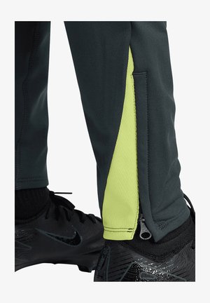 Pantaloni sportivi in tessuto verde scuro con un pannello laterale di un giallo brillante, caratterizzati da un dettaglio con cerniera alla caviglia e da una finitura strutturata.