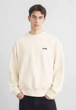 Νέος άνδρας με κοντά σκούρα μαλλιά, φοράει φούτερ ellesse εκρού και μαύρο παντελόνι, στέκεται με τα χέρια στις τσέπες.