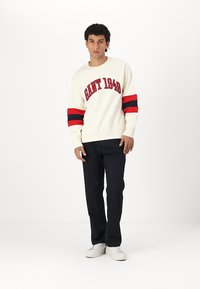 Sweter w odcieniu kremowym z czerwonymi i granatowymi paskami na rękawach, z logo "GANT 1949". W komplecie z ciemnymi spodniami i białymi sneakersami.