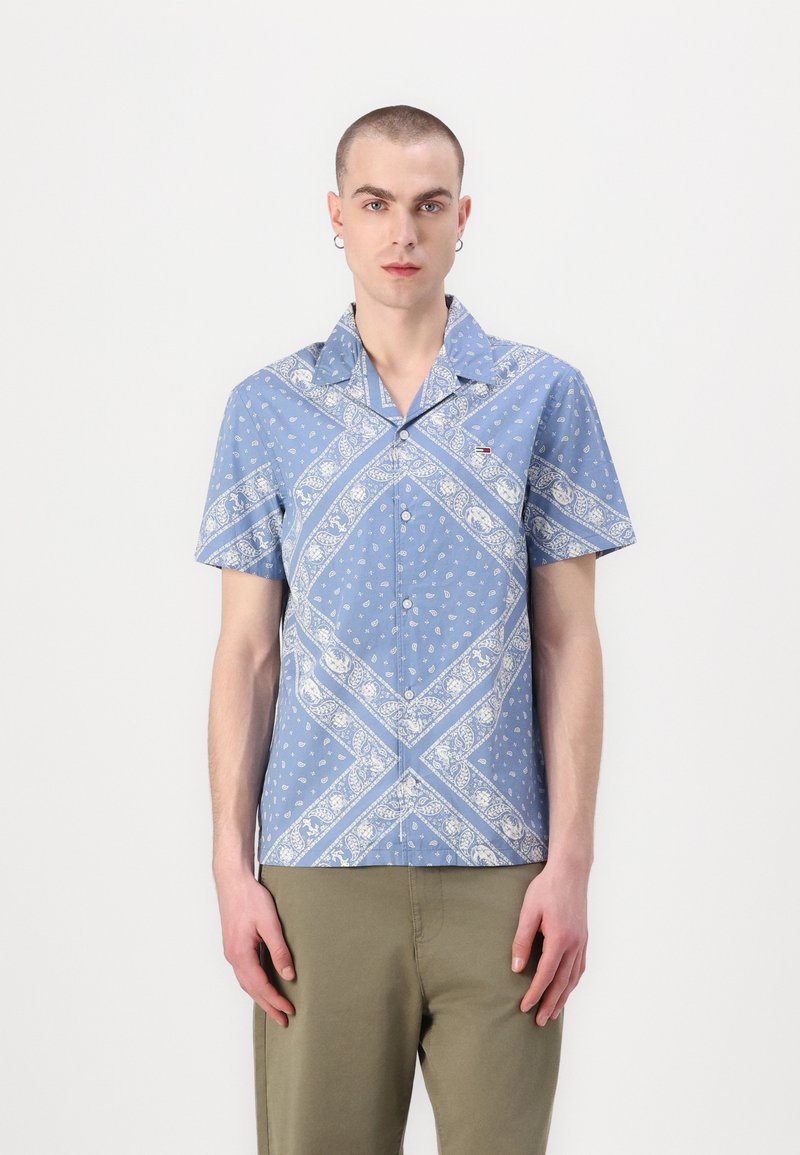 Camicetta azzurro chiaro a maniche corte con motivo paisley bianco e accenti diagonali. Presenta un colletto a camicia e apertura con bottoni.