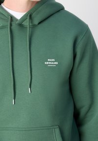 Sweat à capuche vert avec poche avant et texte blanc "MADS NØRGAARD COPENHAGEN" sur la poitrine, porté sur une chemise blanche.