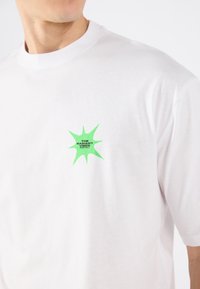 White cotton t-shirt featuring a green graphic with spiky edges and the text "THE RADIANT VIBES FESTIVAL" on the left chest area.