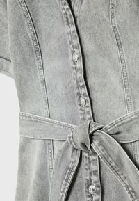 Robe en denim gris clair avec boutons-pression à l'avant et ceinture nouée à la taille, montrant une texture de tissu détaillée et des coutures apparentes.