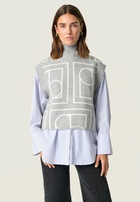 Szürke kötött ujjatlan pulóver magas gallérral, geometriai fehér mintával. Kék és fehér csíkozott ing fölött viselve.