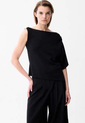Frau trägt ein schwarzes asymmetrisches ärmelloses Oberteil und schwarze plissierte Hosen mit weitem Bein vor weißem Hintergrund.