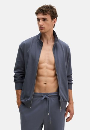 Giovane uomo con capelli ricci che indossa una giacca grigia aperta e pantaloni con coulisse coordinati, mostrando il torso nudo, guardando di lato.