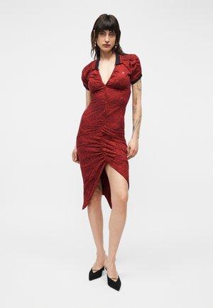 Žena s tmavými vlasmi, oblečená v priliehavej červenej šatke s nariasením, hlbokým výstrihom do V, čiernym golierom, nafúknutými rukávmi, bočným rozparkom a čiernymi špicatými podpätkami.