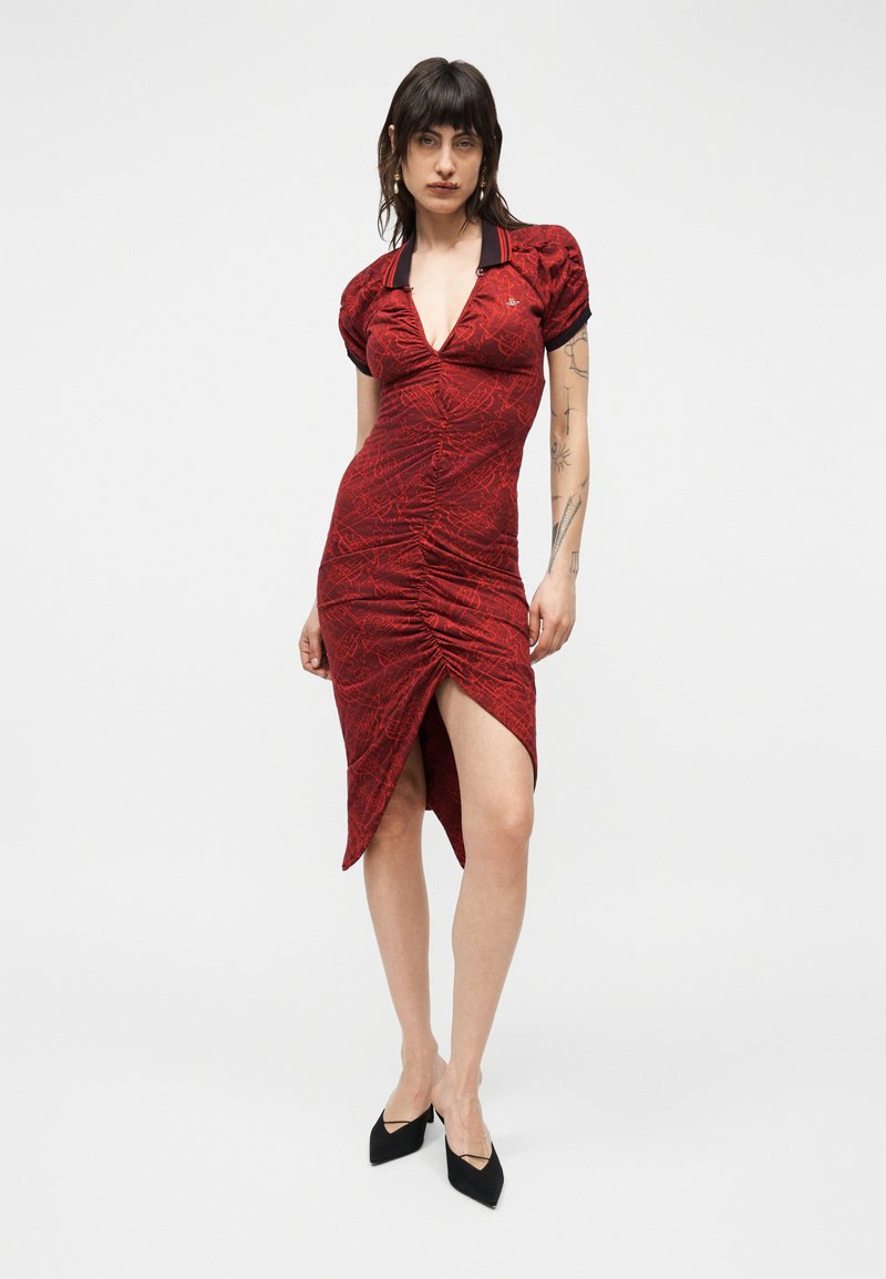 Γυναίκα με σκούρα μαλλιά που φοράει εφαρμοστό κόκκινο φόρεμα με ραφή, βαθύ V ντεκολτέ, μαύρο γιακά, φουσκωτά μανίκια, πλαϊνή σχισμή και μαύρα μυτερά τακούνια.