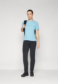 T-shirt atlético azul claro, calças de carga pretas com fechos de correr e sapatos de caminhada pretos. A camisa apresenta um logotipo branco no peito.