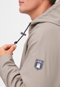Light gray waterproof jacket with adjustable black hood, logo patch on sleeve, and smooth texture. Features a toggle for fit adjustment.