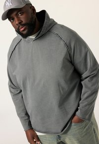 Sudadera con capucha gris, de corte holgado, con costuras en contraste y una textura suave. Combinada con unos jeans de color azul claro.