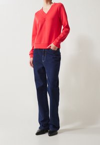 Persona vistiendo una camiseta de manga larga roja con escote en V, jeans rectos de tono azul oscuro y zapatos negros de punta, de pie con una mano en el bolsillo.