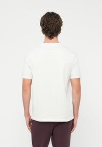 Mężczyzna o kręconych brązowych włosach ubrany jest w prostą białą koszulkę z krótkim rękawem i ciemne spodnie, stoi tyłem do widza na białym tle.