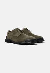 Scarpe in suede verde oliva con suole in gomma nera, caratterizzate da un design con lacci e bordi arrotondati, esposte affiancate su uno sfondo neutro.