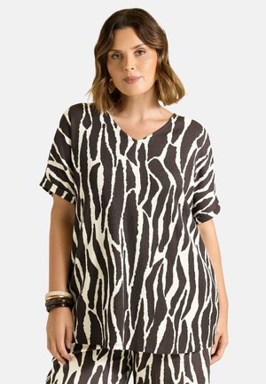Kvinde iført sort-hvid abstrakt zebra print kortærmet top og matchende bukser med armbånd på venstre håndled mod ensfarvet baggrund.