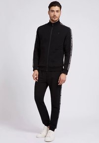 Sort træningssæt med lynlåstrøje og bukser med elastisk talje. Har hvide sidelister med logo. Lavet af glat, behageligt stof.