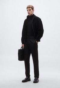 Čierny priliehavý rolák, čierna semišová bomber bunda, šité čierne nohavice a čierne kožené topánky; drží elegantnú čiernu tašku.