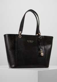 Sac fourre-tout en simili cuir noir avec un motif crocodile texturé, une quincaillerie dorée et un charm logo décoratif. Poignées doubles pour le transport.