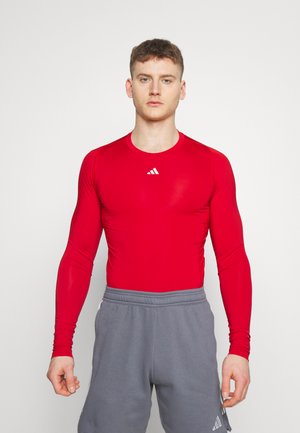 Maglietta sportiva rossa a maniche lunghe con design aderente e scollo tondo, caratterizzata da un piccolo logo bianco sul petto e tessuto liscio e elastico.