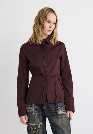 Ξανθιά γυναίκα που φοράει εφαρμοστή σκούρα μωβ πουκάμισο με κουμπιά και λεπτή ζώνη, συνδυασμένο με σκισμένο μπλε τζιν με μεταλλικά τρουκς.