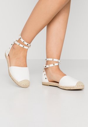 Blote benen in witte leren espadrilles met jute zolen en enkelbandjes versierd met gouden en zilveren piramide-studs.