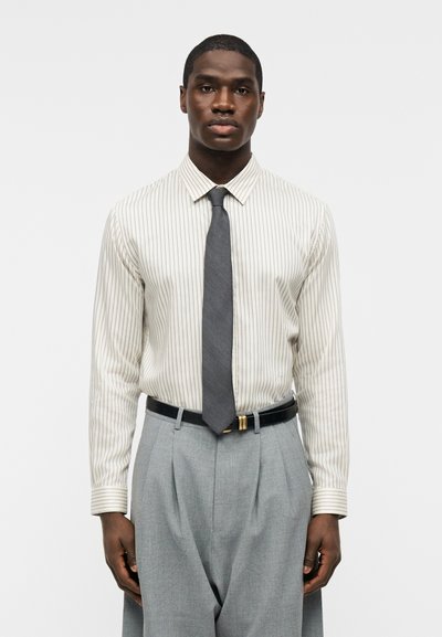 Άνδρας που φοράει λευκό πουκάμισο με λεπτές μαύρες ρίγες, σκούρο γκρι γραβάτα, ανοιχτό γκρι πιετέ παντελόνι και μαύρη ζώνη, στέκεται μπροστά σε απλό φόντο.