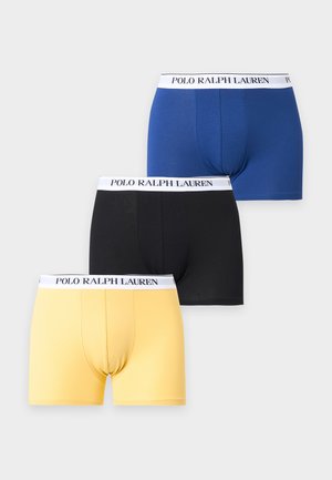 Młody mężczyzna stoi boso, nosząc jasnopomarańczowe bokserki z białym paskiem Polo Ralph Lauren na jednolitym tle.