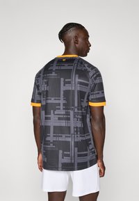 Mand med fletninger iført en sort og grå geometrisk mønstret skjorte med orange kanter, parret med hvide shorts, set fra ryggen.