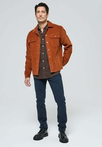 Veste en velours côtelé marron avec deux poches poitrine, associée à une chemise à carreaux foncés et un jean en denim bleu foncé. Complétée par des chaussures à lacets noires.