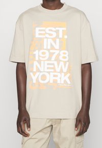 Beige katoenen T-shirt met een grote grafische opdruk in wit en oranje, met de tekst "EST. IN 1978 NEW YORK", een losse pasvorm en korte mouwen.