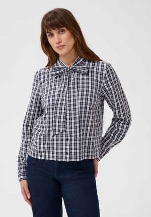 Femme portant un chemisier à carreaux noir et blanc avec un grand nœud noué au col et un jean bleu foncé, debout devant un fond uni.