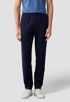 Persona con pantalones deportivos azul oscuro, zapatillas blancas y camiseta azul, de pie frente a un fondo liso gris claro.